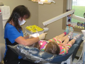 child sitting in dentist chair with assistant holding a tool